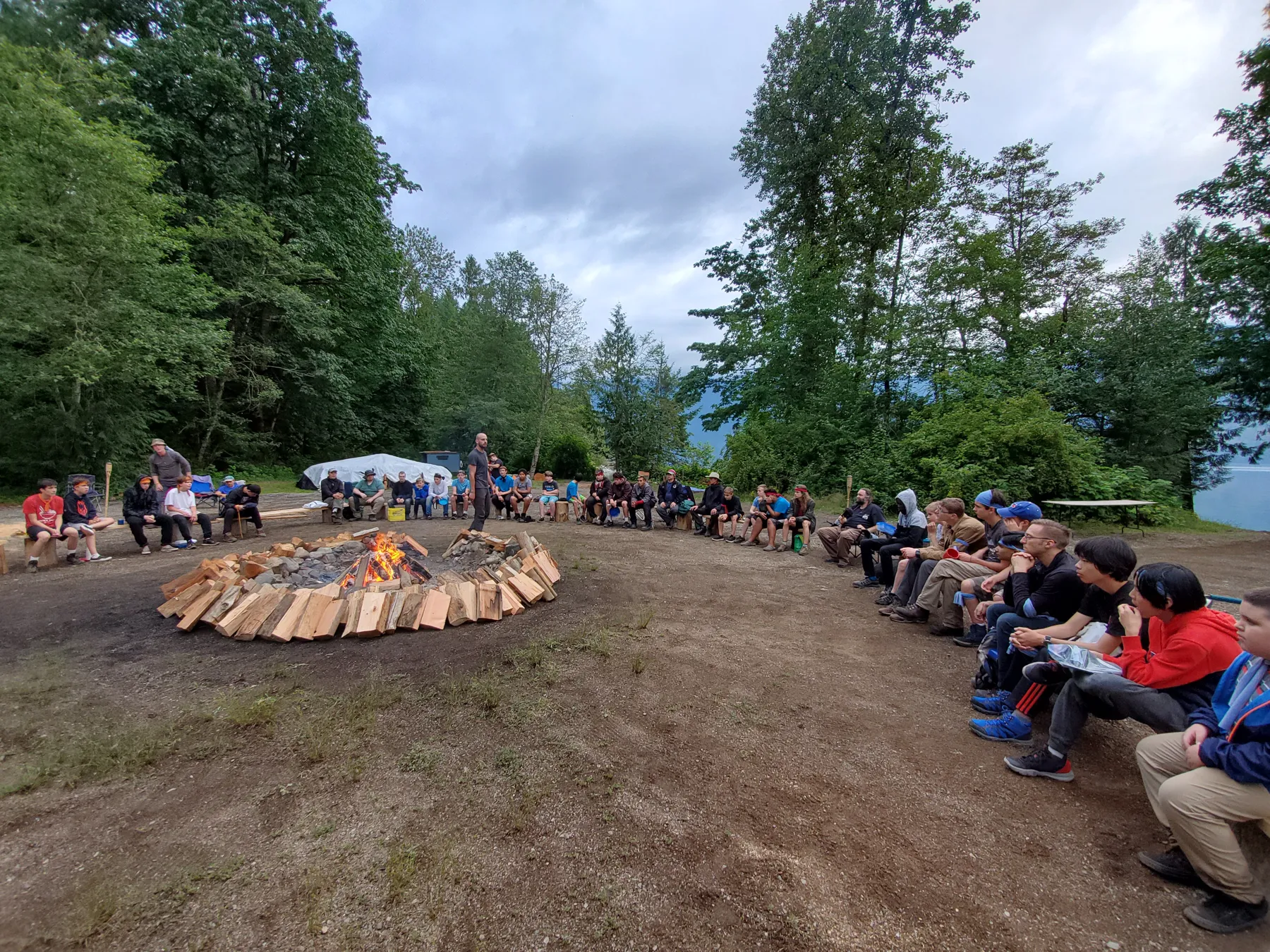 Dusk. Everyone in a circle around the fire pit by the lake, mountains behind