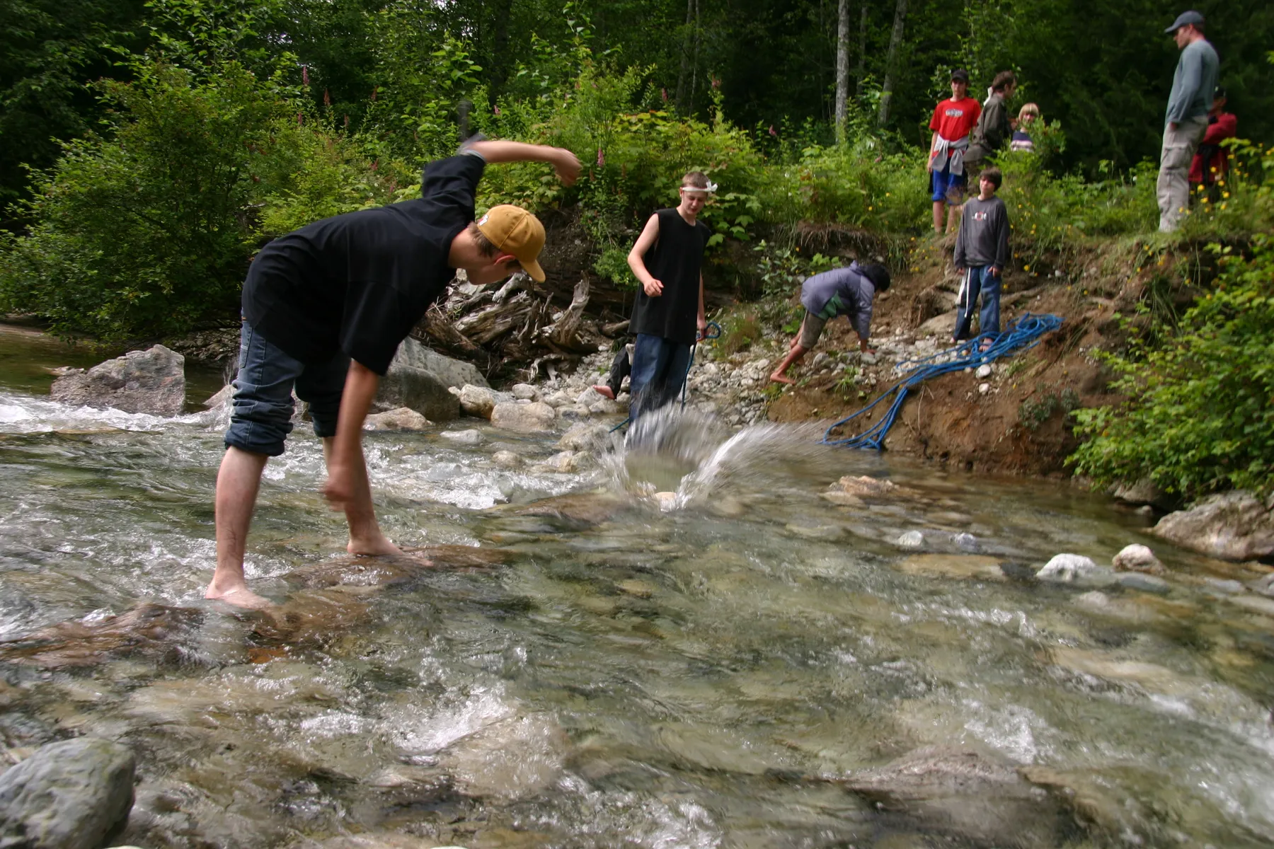 A young man leaps the creek rocks on a Quest, his team behind him